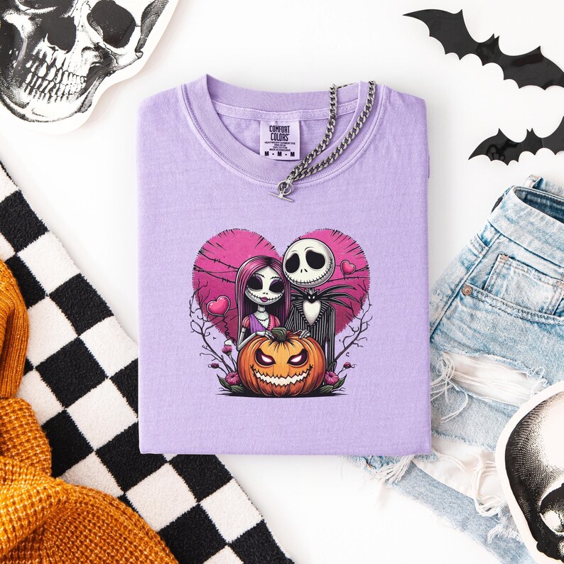 May include: A lilac-colored t-shirt featuring a graphic of Jack and Sally from The Nightmare Before Christmas, set against a pink heart and a jack-o'-lantern. The shirt has a chain necklace and the Comfort Colors brand label.
