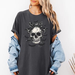 K&ouml;nnte beinhalten: Dunkelgraues T-Shirt mit einem Totenkopf-Motiv, einer Schleife und Flederm&auml;usen. Der Sch&auml;del hat eine schwarz-wei&szlig; gemusterte Schleife. Flederm&auml;use sind um den Sch&auml;del herum verstreut. Das T-Shirt hat hochgekrempelte &Auml;rmel.