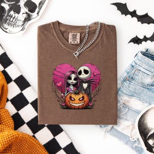 May include: Brown t-shirt featuring a graphic of Jack Skellington and Sally from The Nightmare Before Christmas inside a pink heart, with a carved pumpkin. A silver chain necklace is draped on the shirt. The shirt is folded on a white surface.