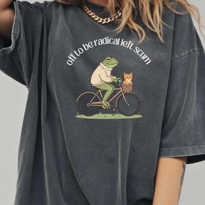 May include: Dark gray t-shirt featuring a whimsical illustration of a frog riding a bicycle with a cat in a basket. The frog wears a sweater, and the text "off to be radical left scum" is arched above the image.