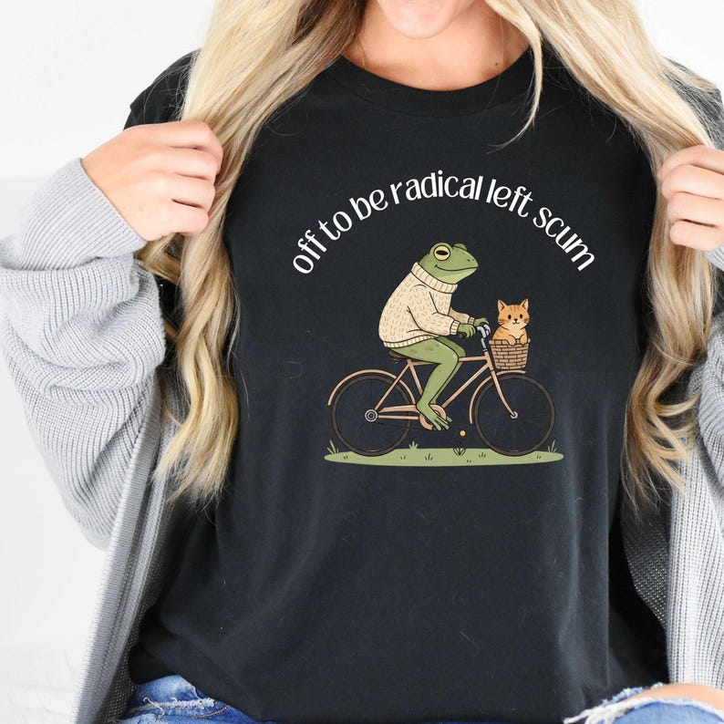 May include: Black t-shirt featuring a whimsical illustration of a frog riding a bicycle with a cat in a basket. The frog wears a sweater, and the text "off to be radical left scum" is arched above the image.