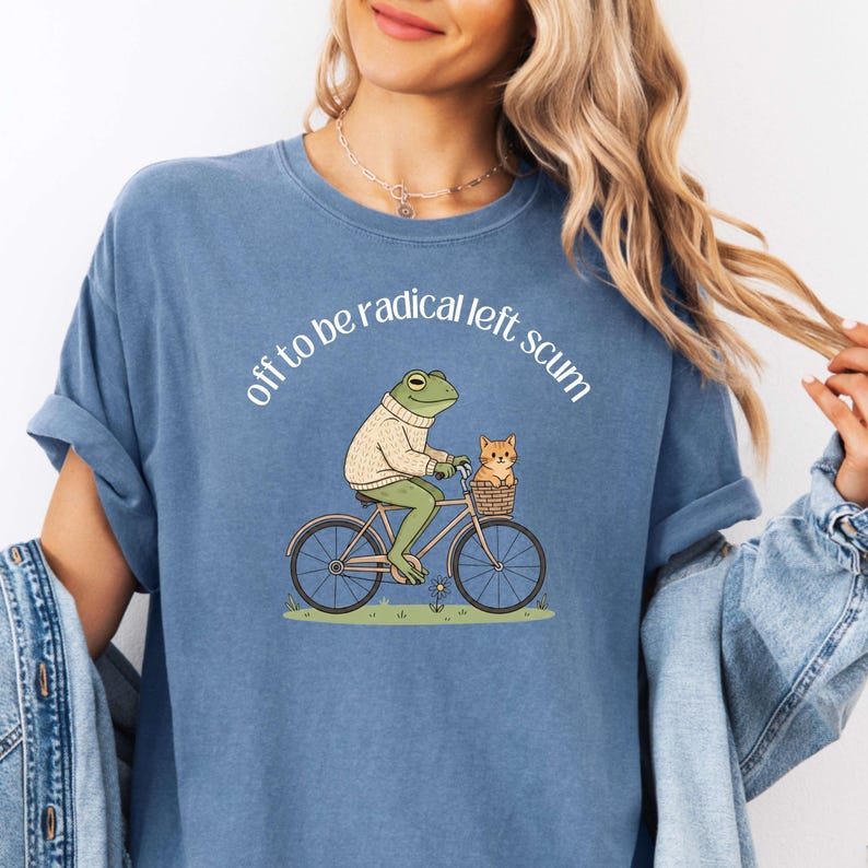 May include: Blue t-shirt with the phrase "off to be radical left scum" in white. The graphic features a frog in a sweater riding a bicycle with a cat in a basket. The design is whimsical and humorous.