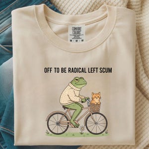 May include: Cream-colored t-shirt with the text "OFF TO BE RADICAL LEFT SCUM." A cartoon frog wearing a sweater rides a bicycle with a cat in a basket. The shirt has a Comfort Colors tag.