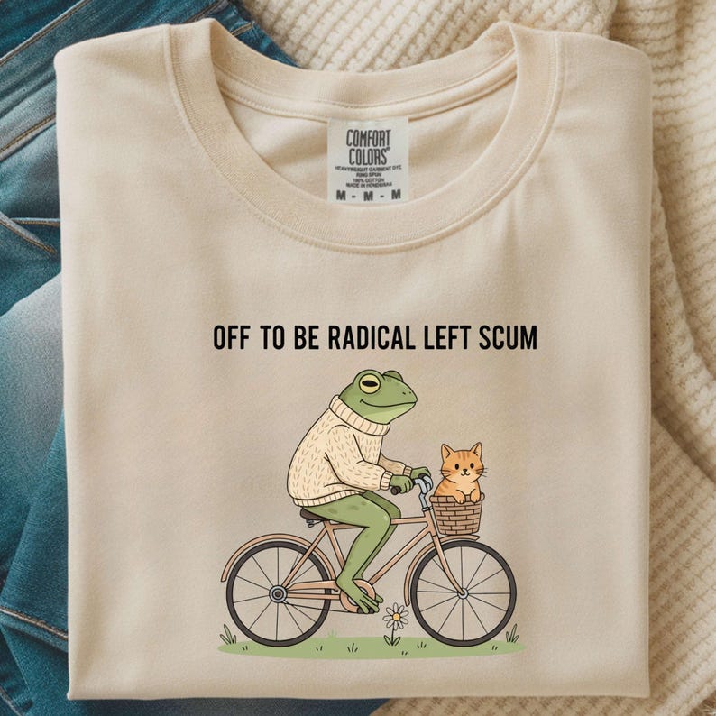 May include: Cream-colored t-shirt with the text "OFF TO BE RADICAL LEFT SCUM." A cartoon frog wearing a sweater rides a bicycle with a cat in a basket. The shirt has a Comfort Colors tag.