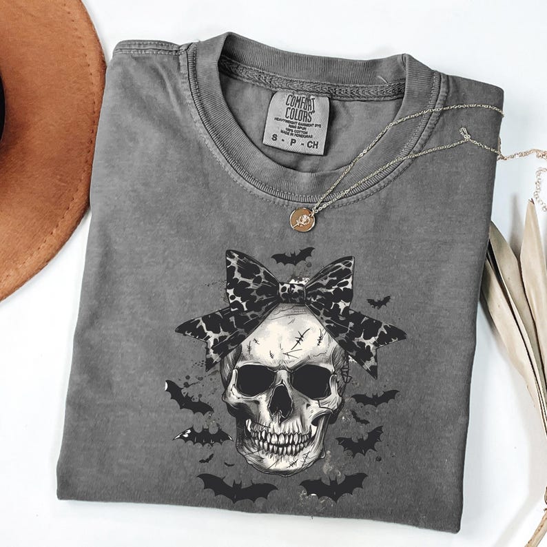 May include: A gray Comfort Colors t-shirt with a skull graphic. The skull has a black and white bow and is surrounded by bats. A silver necklace with a small pendant is draped on the shirt. The shirt has the Comfort Colors logo on the neckline.