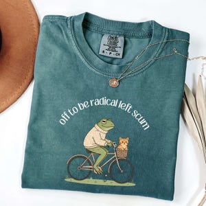 May include: Teal Comfort Colors t-shirt with the phrase "off to be radical left scum" in white. The graphic shows a frog riding a bicycle with a cat in the basket. A gold necklace is draped across the shirt.