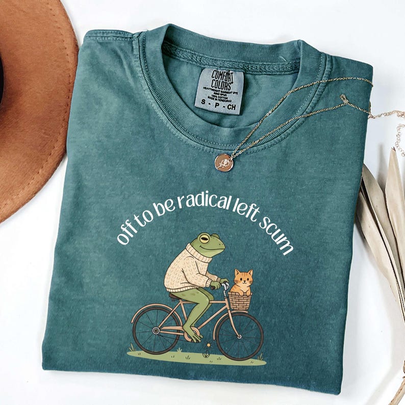 May include: Teal Comfort Colors t-shirt with the phrase "off to be radical left scum" in white. The graphic shows a frog riding a bicycle with a cat in the basket. A gold necklace is draped across the shirt.