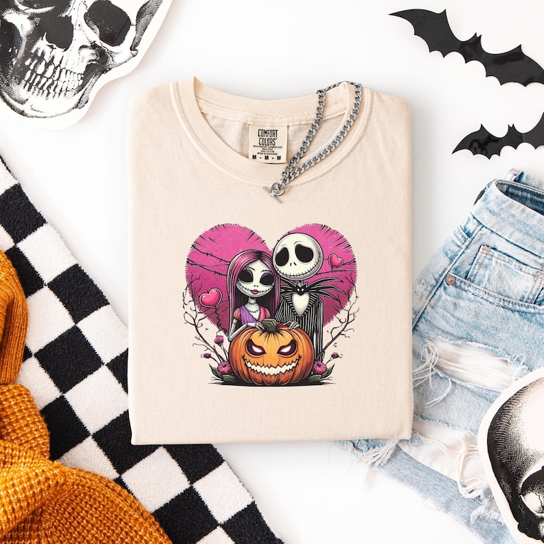 May include: Cream-colored t-shirt featuring a pink heart design with Jack Skellington and Sally from The Nightmare Before Christmas. A carved orange pumpkin sits below them. The shirt has a Comfort Colors tag and a silver chain necklace. Black and white checkered fabric and a skull are visible.