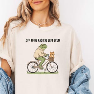 May include: Cream-colored t-shirt with the text "OFF TO BE RADICAL LEFT SCUM." The graphic depicts a frog riding a bicycle with a cat in the basket. The frog wears a sweater. The bicycle is brown and the cat is orange.