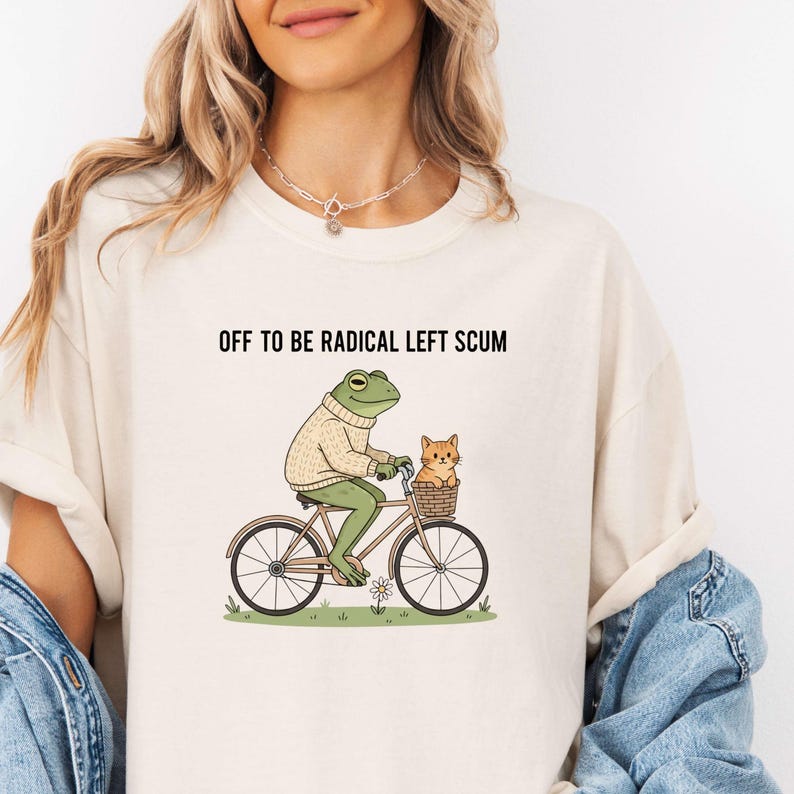 May include: Cream-colored t-shirt with the text "OFF TO BE RADICAL LEFT SCUM." The graphic depicts a frog riding a bicycle with a cat in the basket. The frog wears a sweater. The bicycle is brown and the cat is orange.