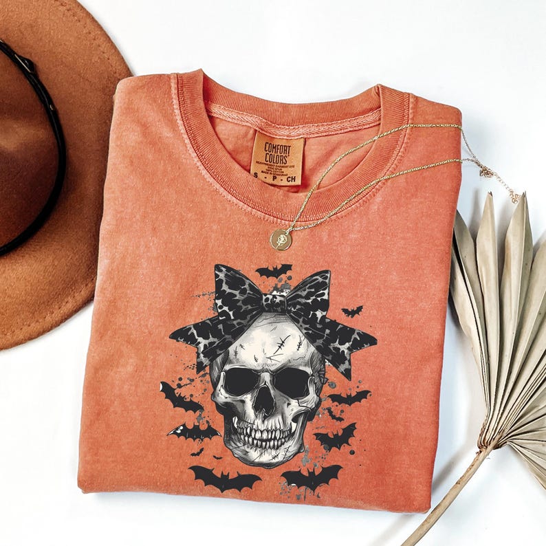 K&ouml;nnte beinhalten: Orangefarbenes Comfort Colors T-Shirt mit einem Totenkopf-Motiv, einer schwarz-wei&szlig;en Schleife und Flederm&auml;usen. Das T-Shirt mit Rundhalsausschnitt hat kurze &Auml;rmel. Eine goldene Halskette mit Anh&auml;nger ist sichtbar. Das T-Shirt ist Gr&ouml;&szlig;e S.