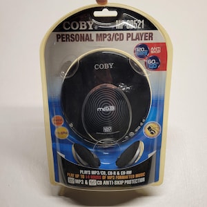 May include: A black Coby MP-CD521 personal MP3/CD player in original blister packaging. The device features a circular design with a central MP3 logo, 120-second anti-skip protection, and includes black headphones. Text includes "Personal MP3/CD Player" and "Plays MP3/CD, CD-R & CD-RW".