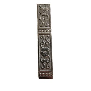 May include: A silver-colored, rectangular metal object with floral and scrollwork designs. The object has a hinged section and a series of small, circular embellishments. Two flowers are visible on the object.