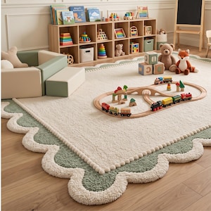 May include: A large, rectangular area rug featuring a cream-colored center with a scalloped border in sage green, accented by a white pom-pom trim. The rug is textured and plush, providing a soft surface for a wooden toy train set and plush toys.