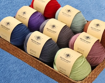 May include: A woven basket filled with nine skeins of Merino wool yarn in various colors, including red, purple, blue, and green. Each skein is labeled with a paper band that reads "Merino wool" and "MMR-212". The basket sits on a blue surface.