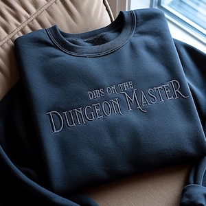 Könnte beinhalten: Ein dunkelblaues Sweatshirt mit dem gestickten Schriftzug "DIBS ON THE DUNGEON MASTER" in einer verzierten Schriftart. Das Sweatshirt liegt gefaltet auf einem beigen Kissen, was seinen lässigen Stil unterstreicht.