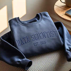 Könnte beinhalten: Ein marineblaues Sweatshirt mit dem gestickten Text "DATA SCIENTIST EST. 2026". Das Sweatshirt liegt gefaltet auf einem gemusterten Stuhl. Eine Kaffeetasse und ein Notizbuch sind im Hintergrund zu sehen.