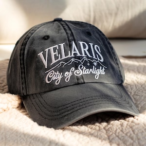 May include: A dark gray baseball cap with the embroidered text "VELARIS City of Starlight" in white. The design includes mountain and star graphics. The cap is made of a soft fabric and has a curved brim.