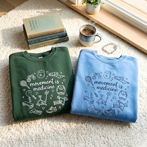 May include: Two crewneck sweatshirts, one in forest green and the other in light blue, are shown on a white rug. Each has the phrase "movement is medicine" with fitness illustrations. A stack of books and a coffee mug are also visible in the image.