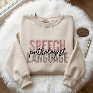Puede incluir: Una sudadera beige con las palabras "SPEECH pathologist LANGUAGE" bordadas en rosa, negro y gris. La sudadera está sobre una superficie blanca y esponjosa, con un bocadillo de madera y un bolígrafo cerca.