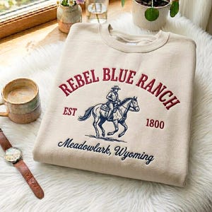 May include: A beige sweatshirt with burgundy embroidered text reading "REBEL BLUE RANCH", featuring a rider on a horse. The text "Meadowlark, Wyoming" is embroidered below. A watch and mug are also visible.