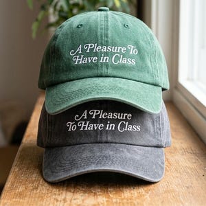May include: Two baseball caps stacked on top of each other. The top cap is green, and the bottom cap is gray. Both caps have the embroidered text "A Pleasure To Have in Class" in white.