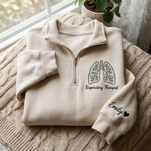 May include: A beige quarter-zip sweatshirt with a black lung graphic and the words "Respiratory Therapist" printed below. The sleeve has the name "Emily" and a heart symbol embroidered in black.