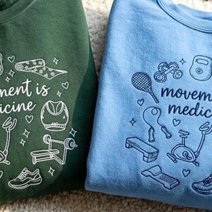May include: Two sweatshirts, one green and one blue, featuring the text "movement is medicine" and fitness-themed illustrations. The designs include a dumbbell, yoga mat, tennis racket, and exercise equipment.
