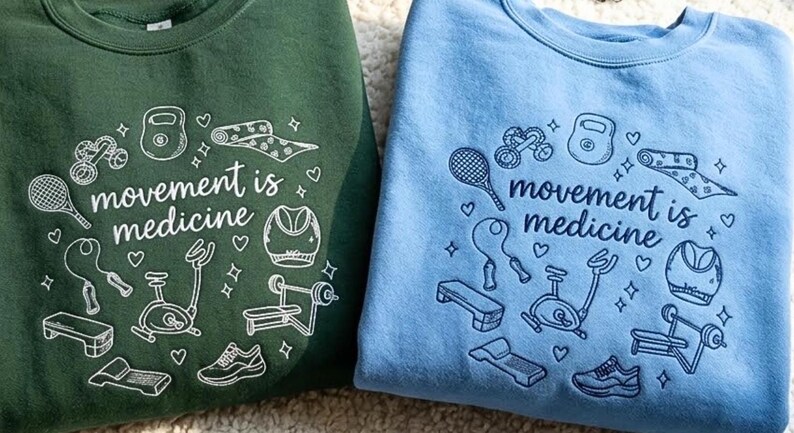 May include: Two sweatshirts, one green and one blue, featuring the text "movement is medicine" and fitness-themed illustrations. The designs include a dumbbell, yoga mat, tennis racket, and exercise equipment.