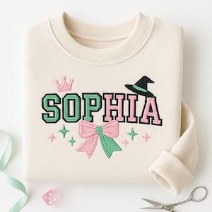 Może przedstawiać: Kremowa bluza z haftowanym imieniem "SOPHIA" w kolorze zielonym i r&oacute;żowym, ozdobiona koroną, kapeluszem czarownicy, kokardą i gwiazdkami. Wz&oacute;r znajduje się na środku przodu odzieży z długim rękawem.