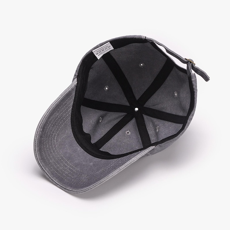May include: A gray baseball cap with a curved brim, viewed from the inside. The cap features a six-panel design with a black interior lining and a fabric strap for adjustment. The cap has a faded, worn appearance.