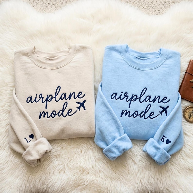 May include: Two crewneck sweatshirts, one beige and one light blue, with the words "airplane mode" embroidered in navy blue script. A small airplane graphic follows the text. The beige sweatshirt has an "L" and a heart on the cuff, and the blue one has a "K" and a heart.