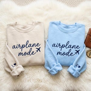 May include: Two crewneck sweatshirts, one beige and one light blue, with the words "airplane mode" embroidered in navy blue script. A small airplane graphic follows the text. The beige sweatshirt has an "L" and heart on the cuff, and the blue one has a "K" and heart.