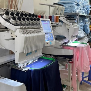 May include: Two white embroidery machines with multiple needles and digital displays. One machine has a blue garment in the hoop, while the other has a pink garment. The machines are in a workshop setting.
