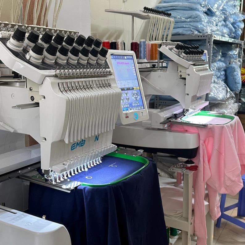 May include: Two white embroidery machines with multiple needles and digital displays. One machine has a blue garment in the hoop, while the other has a pink garment. The machines are in a workshop setting.