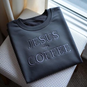 Könnte beinhalten: Dunkelgraues Sweatshirt mit dem gestickten Text "JESUS + COFFEE". Das Rundhals-Sweatshirt liegt gefaltet auf einem hellgrauen Stuhl mit Muster. Das Sweatshirt ist aus einem weichen Material.