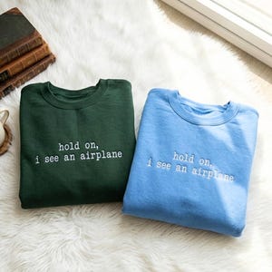 May include: Two crew neck sweatshirts, one forest green and one light blue, are displayed on a white fluffy surface. Each sweatshirt has the phrase "hold on, i see an airplane" embroidered in white.