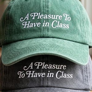 May include: Two baseball caps stacked on top of each other. The top cap is green, and the bottom cap is gray. Both caps have the embroidered text "A Pleasure To Have in Class" in white cursive font.