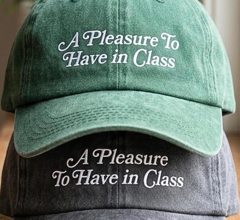May include: Two baseball caps stacked on top of each other. The top cap is green, and the bottom cap is gray. Both caps have the embroidered text "A Pleasure To Have in Class" in white cursive font.