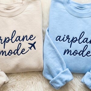 May include: Two sweatshirts, one beige and one light blue, with the words "airplane mode" in navy script and a small airplane graphic. The beige sweatshirt has the letter "L" and a heart on the cuff. The blue sweatshirt has the letter "K" and a heart on the cuff.