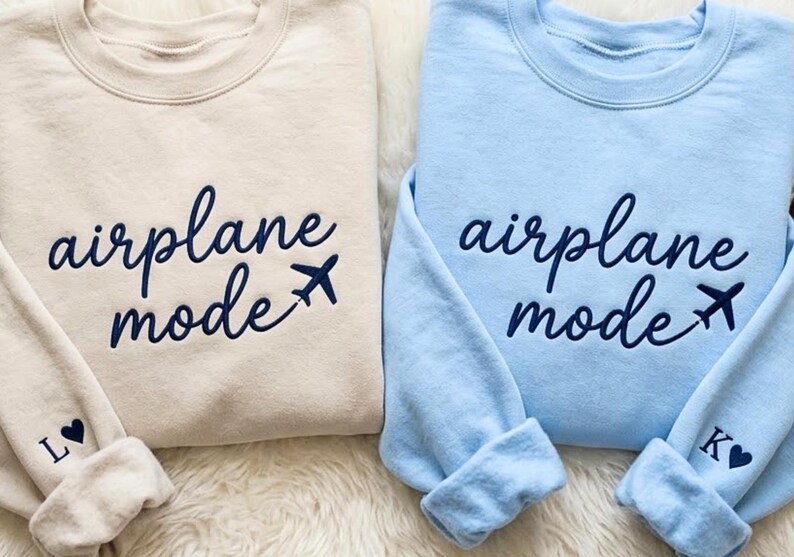 May include: Two sweatshirts, one beige and one light blue, with the words "airplane mode" in navy script and a small airplane graphic. The beige sweatshirt has the letter "L" and a heart on the cuff. The blue sweatshirt has the letter "K" and a heart on the cuff.