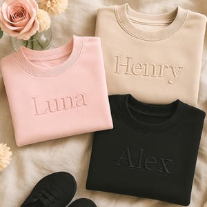 Könnte beinhalten: Drei Rundhals-Sweatshirts in Rosa, Beige und Schwarz, jedes mit einem Namen bestickt: Luna, Henry und Alex. Die Sweatshirts werden auf einer neutral getönten Oberfläche präsentiert, mit einem Paar schwarzer Schuhe im Vordergrund und einer Blumenvase.