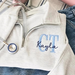 May include: A light gray quarter-zip sweatshirt with the name "Kayla" and the initials "CT" embroidered in light blue. A stethoscope is draped over the sweatshirt, which is lying on a denim surface.
