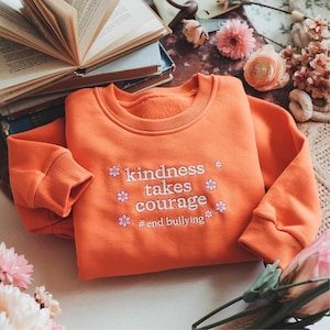 May include: An orange sweatshirt featuring the embroidered phrase "kindness takes courage" and the hashtag "#end bullying". The sweatshirt has a crew neck and long sleeves. Pink and white floral details are included.