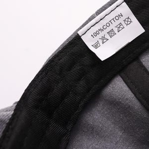 May include: Close-up of a gray baseball cap's interior, showcasing a black sweatband and a white tag. The tag reads "100% COTTON" with care symbols. The cap's fabric appears textured, with visible stitching.