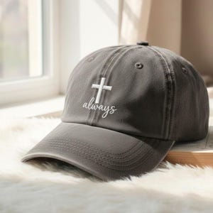 May include: A gray baseball cap with a white embroidered cross and the word "always" in script. The cap has a curved brim and a button on top. The hat is sitting on a fluffy white surface.