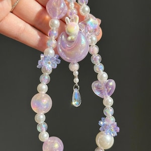 May include: A beaded necklace with iridescent and pastel-colored beads. The necklace features a bunny on a crescent moon, a star, a heart, and flower-shaped beads. A teardrop-shaped charm hangs from the center.