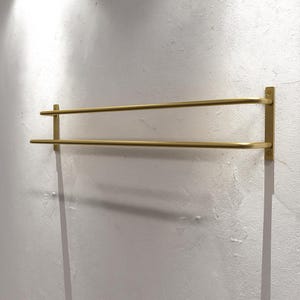 Może przedstawiać: Złoty wieszak na ręczniki z dwoma prętami, zamontowany na białej ścianie. Wieszak ma minimalistyczny design z dwoma równoległymi prętami i prostokątną płytą montażową. Pręty są równomiernie rozmieszczone i wydają się być metalowe.