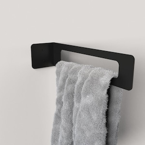 Może przedstawiać: Czarny metalowy wieszak na ręczniki zamontowany na białej ścianie, trzymający szary, puszysty ręcznik. Wieszak ma prostokątny kształt z kwadratową płytą montażową. Ręcznik jest złożony na wieszaku.