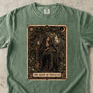 Op de afbeelding: Een saliegroen T-shirt met korte mouwen en een rechthoekige grafische print van een zittende figuur in een donker gewaad, omringd door gebladerte. De tekst THE QUEEN OF PENTACLES staat onderaan de illustratie, die een zon en maan bevat.